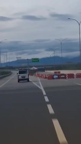 A white van is seen driving on a highway. The van is traveling in the right lane, and there are other cars in the distance. The highway has multiple lanes, and there are streetlights and signs visible. The sky is cloudy, and there are mountains in the background. The van appears to be an ambulance, with red and white markings on its side. There are orange traffic cones and barriers on the right side of the highway. There is also a tent set up on the right side of the highway.