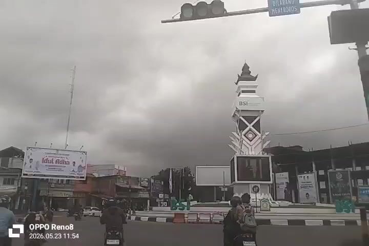 The video depicts a street scene in what appears to be a town square or roundabout. Several people are riding motorbikes on the road, moving across the frame. In the background, there are buildings of varying heights, indicative of a commercial area. A tall, white, ornate structure resembling a clock tower stands in the center of the roundabout, with the letters "BSI" visible on its side. There are also large banners or billboards displayed, one of which advertises "Idul Adha," indicating a Muslim holiday. The sky is overcast with heavy, grey clouds, suggesting a potentially rainy or gloomy weather. The video is taken from a moving vehicle or person, creating a dynamic perspective of the scene. The timestamp in the lower-left corner indicates the video was recorded on May 23, 2026.