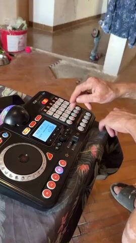 The video shows a close-up of a person's hands operating a DJ controller. The controller is black with a variety of buttons and a small screen displaying text. The person's fingers are seen pressing different colored buttons on the controller. The background is slightly out of focus but appears to be an indoor setting with wooden flooring and some furniture visible.