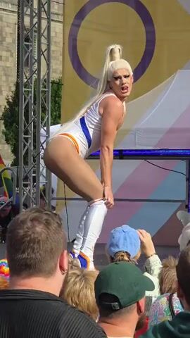 A drag performer with a blonde ponytail and white bodysuit is on a stage. They are bending over and adjusting their outfit. The performer has white makeup and is wearing white boots. Behind the performer, there is a large banner with a yellow and purple circle and a striped pattern. There are people in the audience watching the performance.