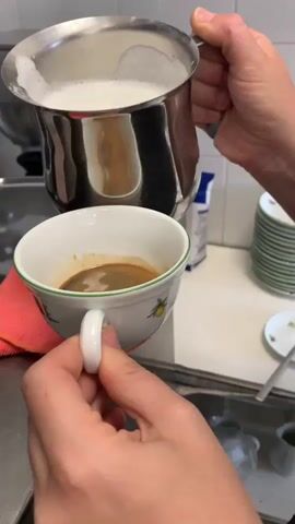 A close-up shot shows a person's hands preparing a coffee drink. The person is holding a white ceramic mug with a green rim and a fruit design, filled with dark coffee and topped with a layer of frothy white milk. In their other hand, they hold a stainless steel pitcher containing more frothed milk, which they are pouring into the mug. The milk is creating a swirling pattern on the surface of the coffee. In the background, a stainless steel sink, a coffee machine, and stacked white plates with green rims are visible. The scene appears to be in a kitchen or cafe setting.