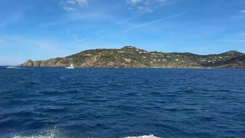 The video shows a view from a boat moving across the ocean. In the foreground, the blue water is choppy, with white spray visible on the left side of the frame, indicating the boat's movement. In the middle ground, a white sailboat is sailing on the water. In the background, a hilly, green island is visible, dotted with houses and a lighthouse or tower on the highest peak. The sky is blue with scattered white clouds and contrails. The overall scene depicts a sunny day at sea with a view of a coastal landscape.