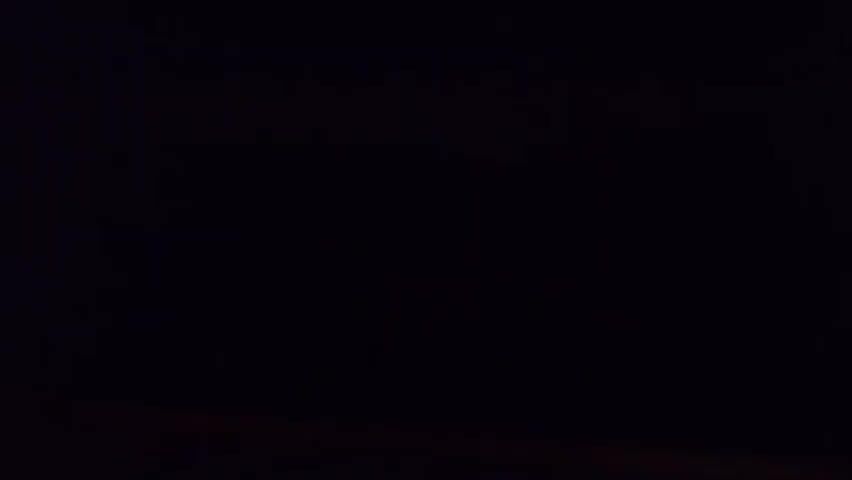 The video opens with a completely black frame. After a moment, a faint reddish light appears in the upper right quadrant of the frame. As the video progresses, faint lights, presumably from a distant settlement or city, become visible near the bottom of the frame, creating a horizontal line of illumination against the dark background. The lights appear scattered and somewhat indistinct, suggesting distance and possibly atmospheric conditions. The overall scene is dark and captures the distant glow of artificial lights against a night sky.