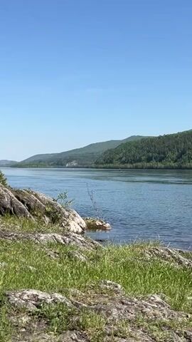 The video presents a serene landscape featuring a wide body of water, likely a river or lake, under a clear blue sky. In the foreground, a rocky shoreline is partially covered with vibrant green grass. A small, sparse tree or bush grows from the water's edge amidst the rocks.

Across the water, a long stretch of verdant, forested hills or mountains dominates the horizon, indicating a natural, perhaps mountainous, setting. The water itself is calm with only slight ripples, reflecting the brightness of the sky. In the far distance on the water, a tiny, dark shape is visible, possibly a small boat or a distant figure. Overhead, subtle power lines or cables stretch across the sky, suggesting some human presence in the broader area despite the otherwise natural scene. The lighting indicates it is bright daylight, likely morning or afternoon, with clear weather. No specific activity is prominently featured, contributing to a sense of tranquility. There is no visible text. The location is Sliznevo, Russia.