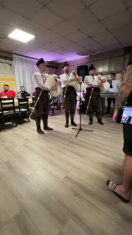 Three men in traditional attire play bagpipes indoors. They stand in the center of a room with light wooden flooring. The men wear white shirts with embroidered patterns, dark brown trousers, and dark hats. Each holds a light-colored bagpipe. In the background, several individuals are seated at tables, and to the right, another man stands near a keyboard. A person on the far right, partially visible, is holding a phone, seemingly recording the performance. The room has light-colored walls, and the ceiling features a rectangular light fixture and purple lighting.
