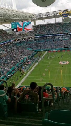 The video shows a football game being played in a stadium from a high-angle perspective. The stadium is full of people, and a football field can be seen below. The Hard Rock Stadium sign is visible. Several people are sitting in the seats in front of the camera, watching the game.  The players on the field are spread out, engaged in active gameplay. The overhead screen shows the game score and a dolphin image. The weather is bright, with some clouds in the sky.