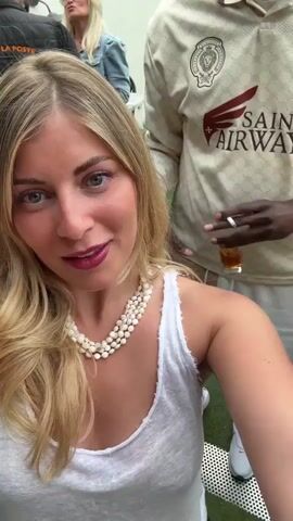 A blonde woman with a pearl necklace and a white tank top is taking a selfie. A black man with a white cap and sunglasses puts his arm around her and makes a peace sign. They both smile and laugh at the camera. The background is an outdoor setting, possibly a restaurant or patio, with people sitting at tables. The man is wearing a cream-colored shirt with a logo and the words "SAINT AIRWAYS" on it. The woman has dark lipstick and purple eyeshadow.