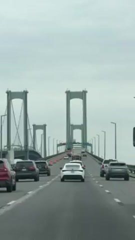 The video shows a car driving on a bridge with multiple lanes, moving with other vehicles. The bridge has tall, arch-shaped support structures, and there are traffic lights overhead. The sky is overcast.