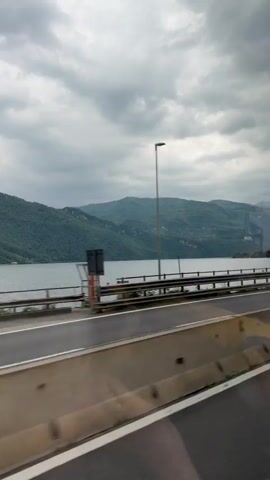 The video shows a view from a moving vehicle, likely a car or bus, traveling along a highway next to a lake. The highway has a concrete barrier and a metal guardrail along the lake side. Beyond the guardrail is a large lake surrounded by green, hilly terrain. The sky is overcast with gray clouds. The road surface is asphalt with painted white lane markings. The perspective suggests the video was taken from the passenger side of the vehicle. The scene remains consistent throughout the duration of the video.