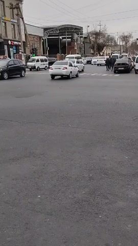 The video shows a busy intersection in a city. There are many cars and vans moving through the intersection. People are walking on the sidewalks and crossing the street. A person on a bicycle with a basket in the front rides across the intersection. There are buildings on the sides of the road, including one that has a sign in a foreign language. The sky is overcast and the weather appears to be cloudy.
