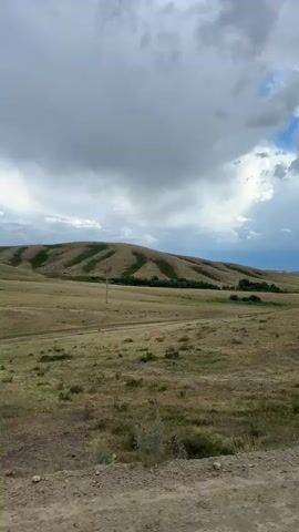 This media file depicts a rolling, arid landscape under a dramatic, cloudy sky. The foreground is a dusty, unpaved road with tire tracks, suggesting recent vehicle activity. Beyond the road, the terrain transitions to dry, yellowed grasses and scattered low-lying shrubs. In the mid-ground, a series of rounded hills dominate the scene, with distinct bands of darker green vegetation creating a striped pattern across their slopes. A solitary utility pole stands in the middle distance, breaking the natural uniformity. The background reveals more distant hills fading into the horizon.

The weather appears to be transitioning, with heavy, dark grey clouds dominating the sky, interspersed with patches of bright blue and shafts of sunlight breaking through. This suggests a possibility of rain or a recent storm. The time of day appears to be late afternoon or early evening, given the quality of the light.

The overall mood is one of vastness and natural beauty, with a touch of ruggedness due to the arid conditions and unpaved road. The landscape evokes a sense of open space and possibly a remote location. There are no visible people or man-made objects other than the utility pole and the road.

The location context provided is Bakty, Kazakhstan, which aligns with the arid, steppe-like environment shown in the video.