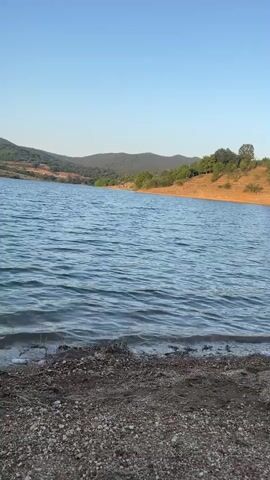 This is a vertical video clip of a tranquil lake scene at Domlyan, Bulgaria. The camera is positioned low to the ground, at the edge of the water. In the foreground, there is a stony shoreline with scattered dry grass and twigs. The lake water is a vibrant blue, with gentle ripples creating a shimmering effect under the clear sky.

In the background, rolling hills covered in lush green trees and vegetation stretch across the horizon. The hills are bathed in the warm, soft light of the late afternoon or early evening sun, casting subtle shadows and highlighting the contours of the landscape. The sky above is a pale, clear blue, indicating fair weather. There is no text visible in the clip.

The overall mood of the video is peaceful and serene, evoking a sense of calm and natural beauty. The lack of any human presence or activity further enhances this feeling of solitude and quietude. The scene suggests a moment of undisturbed nature, possibly at a time of day when the light is most flattering and the environment is at its most picturesque.