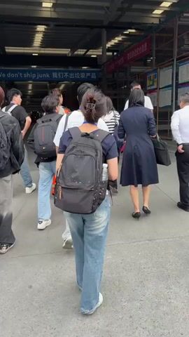 A group of people walk into a warehouse. There are about 12 people, mostly young adults in casual wear and backpacks. Two women are dressed more formally, one in a navy dress and one in a striped shirt. Two men wear white dress shirts and black pants. One of these men speaks to the woman in the navy dress. A sign over the entrance reads “We don’t ship junk.” The warehouse interior is visible in the background.