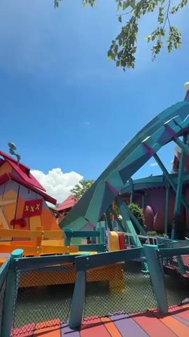 The video shows a water ride at an amusement park on a sunny day.  The ride features colorful, whimsical structures in yellow, orange, teal and purple.  There is a boat on a track climbing an aqua-colored hill. A small red and yellow building with a flag marked "XXX" sits next to the track. The ride then enters a water channel with large orange sphere-shaped obstacles.  The water is clear and blue, and the sky is a bright blue with some white clouds. Trees surround the area. There's black netting on the edge of the water, obscuring part of the view.