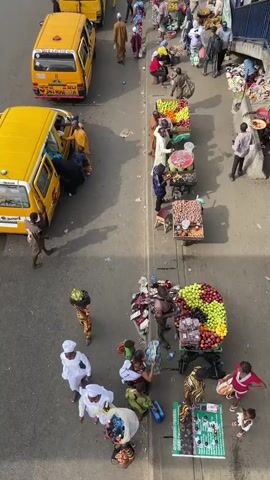 The video shows a high-angle shot of a busy street scene in what appears to be Nigeria. Several yellow vans are parked on the side of the road, with people getting in and out. Along the sidewalk, there are vendors selling fruits and other goods from carts. Many people are walking by, some carrying goods on their heads. The overall environment is bustling with activity.