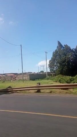 The video, shot from inside a moving vehicle, captures a view of a roadside scene. The day is sunny with a blue sky and scattered clouds. The road in the foreground has yellow lane markings.

On the left side of the road, there's a grassy area where some animals, possibly goats or sheep, are grazing. Behind them is a weathered, possibly concrete, wall with what appears to be graffiti or faded signage. A large metal structure stands near the road. It appears to be a signpost structure without the actual sign.

As the video progresses, a white truck moves rapidly past the camera, followed by a blurred silver vehicle. The view shifts to reveal more of the surrounding area. There are small buildings or structures visible in the background, and a sign with a phone number is seen on one of the buildings. Overhead power lines and utility poles dot the landscape. Some people can be seen on the side of the road. The environment appears somewhat rural or peri-urban.