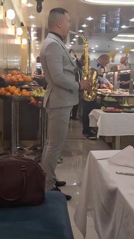 A man in a gray suit plays a golden saxophone in a restaurant. He is facing away from the camera and towards a food buffet table where people are selecting food. To the left of the man is a table with fruit, and to his right is a table with a white tablecloth and a napkin. A brown leather bag sits on a blue seat in the foreground. The restaurant has a mirrored ceiling and is well-lit. The man appears to be performing for the restaurant patrons.