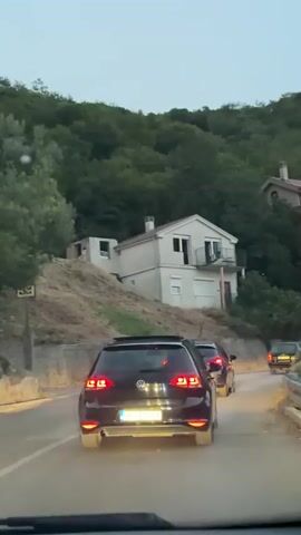 The video shows a rear view of a black Volkswagen Golf driving on a road in a hilly, possibly mountainous area. Several other cars are visible ahead, also driving on the road. On the left side of the road, there are houses built into the hillside, one of which is white with a balcony and another which is larger and beige with multiple balconies. The area is densely covered with green trees. The sky is overcast and appears to be dusk or early evening. The video is shot from inside another vehicle.