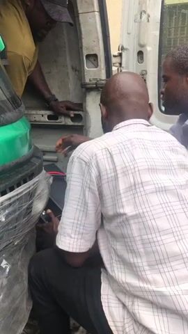 Three men are gathered around the open rear door of a white van. One man, wearing a yellow shirt and a dark cap, is partially visible on the left, resting his hands on the interior of the van. In the center, a man wearing a white and black checkered shirt is kneeling and appears to be working on a black rectangular object with red wires. He is holding a tool in his right hand. To the right of the kneeling man, another man in a white collared shirt is standing and looking towards the black object. The environment appears to be outdoors, with some greenery visible on the left side of the frame and what looks like a solar panel or similar flat object with a grid pattern in the background on the right. The ground is uneven and appears to be made of dirt or gravel.