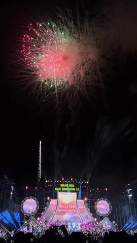 The video captures a vibrant night-time celebration in Măng Đen, Vietnam, featuring a spectacular fireworks display above a large, illuminated stage. The time of day is clearly night, with the dark sky providing a stark contrast for the bright pyrotechnics.

The central focus is the elaborate stage, which is designed with multiple large screens, bright spotlights, and various decorative elements. On the main screen, the text "MĂNG ĐEN KHÁT VỌNG VƯƠN XA" (Măng Đen, Aspiration to Reach Far) is prominently displayed in a bold, stylized font, indicating the event's theme or location. This text is also visible on a smaller screen on the lower part of the stage. Performers, dressed in bright outfits, are visible on the stage, though their specific actions are not detailed due to the focus on the fireworks.

Above the stage, a continuous fireworks show unfolds. The fireworks launch from behind the stage and burst into various patterns and colors against the black sky. Red and golden bursts are particularly noticeable, creating grand, cloud-like explosions and long, sparkling trails. The sound of the fireworks is audible, adding to the dynamic atmosphere.

Below the stage, a large audience is visible as dark silhouettes, indicating a significant turnout for the event. The bright stage lights illuminate the area, adding to the festive and exciting ambiance. The overall scene suggests a grand public celebration, possibly a concert or a festival, marked by a sense of pride and aspiration, as hinted by the visible text.