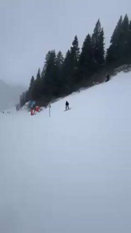 The video shows a snowy ski slope with several skiers and snowboarders heading downhill. The weather is overcast, and the mountains in the background are partially obscured by clouds. There are pine trees lining the side of the slope and a red safety net to the left. The snow is predominantly white with visible tracks from previous skiers and snowboarders.