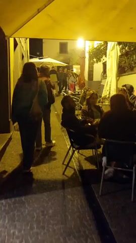 The camera pans slowly through a narrow, cobblestone-paved street scene at night. Illumination comes from overhead lights, casting a yellowish glow. The street is lined with buildings, some with outdoor seating areas occupied by people. Several individuals are walking away from the camera down the street. At one seating area, a few people are seated at tables, some smoking. A sign indicating an ATM is visible on one of the buildings.