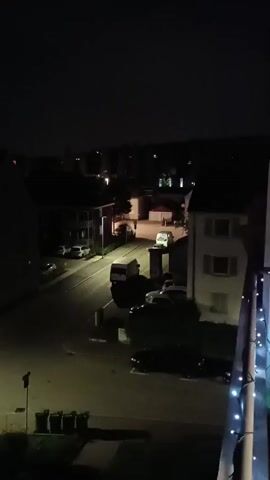 The video shows an elevated, nighttime view of a street in a residential area. The scene is dark with streetlights illuminating sections of the road and nearby buildings. Several parked cars and a van are visible on the street. In the distance, other buildings and lights can be seen. The video appears to be shot from a balcony, with the railing visible in the foreground, adorned with small decorative lights. A person can be briefly seen walking in the distance.