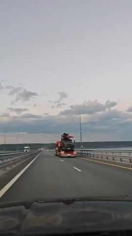 A car is driving on a bridge over a body of water during dusk. The sky is overcast with clouds, and the sun is setting, casting a soft light on the scene. The road is paved and has multiple lanes. On the left side of the road, there is a guardrail separating the road from the water. On the right side of the road, there are streetlights and more guardrails. A large truck carrying a heavy load is visible in the distance on the right lane. The interior of the car is partially visible in the foreground, showing the dashboard and the windshield. The camera is mounted inside the car, giving a first-person perspective of the drive.