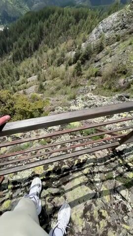 From a high vantage point, the video captures a scenic landscape of rolling hills covered in lush green forests under a bright blue sky dotted with fluffy white clouds. The viewer is positioned on a rocky outcrop protected by a metal railing. The camera pans upwards to reveal the expansive panorama. Below, the landscape slopes steeply down, showcasing the depth and vastness of the mountainous terrain. A small village can be spotted nestled among the trees in the distance. The overall impression is one of natural beauty and tranquility. A person is standing behind the metal railing.