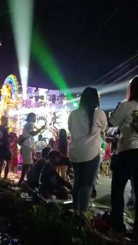 The video shows a nighttime outdoor gathering with a large illuminated stage featuring multiple speakers and lights, and a crowd of people. Some individuals are standing and watching the stage, while others are seated on the ground. The lighting is dynamic, with bright and colorful lights, including green and purple, flashing across the scene. There are power lines visible overhead. The overall atmosphere appears to be that of a public event or festival.