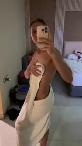 A woman stands in a bedroom, facing a mirror and taking a selfie with her phone. She is partially covered by a white towel, holding it to cover her chest. The room contains a bed, a gray chair with orange pillows, a small black table, and tiled floors. The walls are painted in neutral colors. The lighting is soft, illuminating the scene. The woman's phone obscures her face in the reflection.