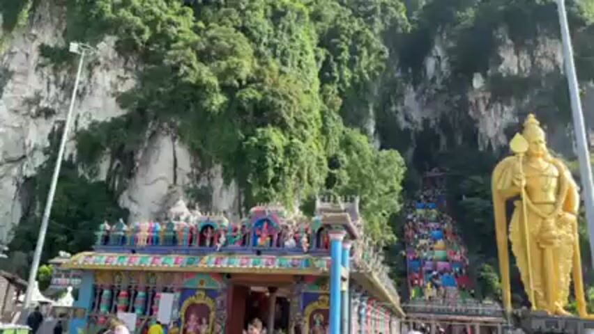 The video showcases the Batu Caves in Malaysia, a Hindu shrine and popular tourist attraction. The view is from ground level, facing a towering gold statue of Lord Murugan. Behind the statue, a steep, multicolored staircase leads up to the cave entrance in the limestone hill. The hillside is covered in green vegetation. To the left and right of the staircase are colorful temple structures. A crowd of people, including tourists and locals, are seen walking around the area, some ascending the staircase. The lighting suggests a bright, sunny day. The video provides a clear and wide shot of the temple complex and its surroundings.