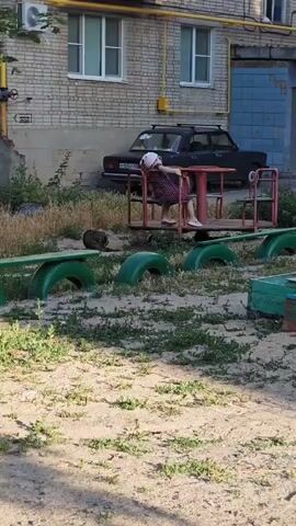 This video shows a young girl in a polka-dot dress sitting on a merry-go-round in a playground. The merry-go-round appears to be made of repurposed materials, with old tires painted green used as part of the playground's features. Behind the playground, a dark-colored car is parked in front of a brick building. The building has several windows and an air conditioning unit is visible on the left side. There is also a yellow pipe running down the side of the building. The ground is covered in sand and patchy grass, suggesting an outdoor setting in a somewhat unkempt urban or suburban area. A cat can be seen near the merry-go-round. The lighting suggests it is daytime, likely late afternoon or early evening due to the warm, somewhat long shadows. The overall atmosphere appears to be relaxed and casual, with the child seemingly enjoying herself on the playground equipment. There is text visible on a metal sign on the building in the background, but it is not clearly legible. The license plate of the car is also visible but unreadable. The location is identified as Belaya Kalitva, Russia.