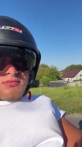 A man wearing a black LS2 helmet with a dark visor and a white t-shirt is riding on a vehicle, possibly a motorcycle or scooter, as indicated by the reflection in his visor. He has a light-colored beard and is looking forward. In the background, there is a rural setting with houses, trees, and a clear blue sky. Another person wearing a black helmet is visible behind him, to the right. The video appears to be shot from a first-person perspective, possibly by a passenger or from the perspective of the rider.