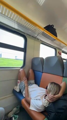 The video shows a person lying down on a train seat with their legs propped up. They are wearing a white t-shirt, denim shorts, and white socks. Their head is resting on their arm. The train appears to be moving, as indicated by the blurred green landscape visible through the window. The seats are upholstered in blue, orange, and gray. A black backpack is visible on the overhead storage rack. Two pairs of sneakers are on the floor near the person's feet.