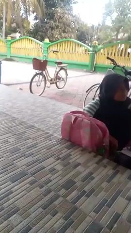 A person wearing a black hijab and a striped shirt kneels on a tiled floor, rummaging through a pink and white patterned bag. Next to them is a dark-colored bag. In the background, two bicycles are parked, and a green and yellow fence encloses an area with trees. To the right, a light blue scooter is visible, along with a sign board. Another person in a blue skirt walks past in the foreground.