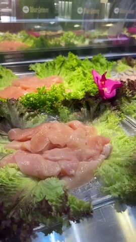 The media file shows a close-up view of a food display in what appears to be a restaurant or buffet. The display features fresh ingredients, primarily raw chicken and various types of lettuce, presented on ice. There are also some cooked meats, possibly duck, being handled with black tongs in the foreground. Bright pink orchids are strategically placed among the food items, adding a decorative touch.

In the background, glass cabinets are visible, hinting at a larger display area. Some green signage with the word "Warsteiner" is visible on these cabinets, which is a German brewery brand, suggesting the location is likely in Germany, specifically Herford as provided in the context. The lighting appears to be artificial, typical of indoor food displays, and there are no indications of time of day or weather conditions. The overall impression is one of freshness and a well-presented selection of ingredients. The focus is entirely on the food and its presentation, with no people visible in the frame.