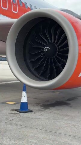 A close-up shot shows an orange and white airplane engine, with a blue and white traffic cone placed beneath it. The engine has a large, circular opening with visible fan blades inside. The plane is parked on a concrete surface with some yellow markings. The sky is partly cloudy.