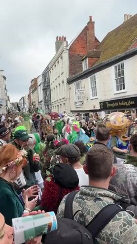The video captures a street parade with numerous people dressed in costumes, mostly green and nature-themed, moving down a narrow street lined with multi-story buildings. The crowd is dense, with spectators on both sides and in between the costumed participants. The buildings are a mix of brick and white-painted facades, and signage for shops like "Roberts Curios" and "Through the looking glass" are visible.  The sky is overcast.  Many spectators have drinks in their hands.