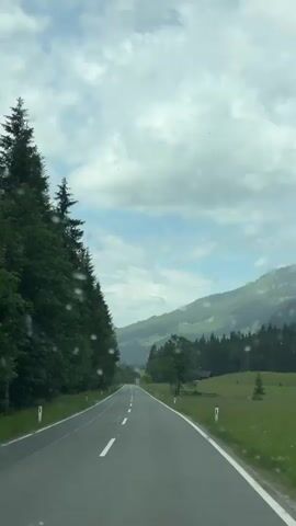 This is a vertical video shot from inside a vehicle, looking out through a rain-streaked windshield. The view is of a paved road winding through a lush green mountain landscape. On the left side of the road, a dense forest of tall, dark green coniferous trees lines the roadside. On the right, a grassy field slopes gently upwards towards more forested hills. The road itself is asphalt with white lane markings.

In the distance, the road continues to curve and ascend into a valley, with more layers of green mountains visible, some shrouded in mist. The sky above is overcast with dramatic, puffy white clouds interspersed with patches of blue sky. The presence of raindrops on the windshield is a prominent feature of the video, indicating it is raining or has recently rained. The lighting suggests it is daytime, likely late morning or afternoon.

There are no people or specific objects that are clearly identifiable beyond the natural elements and the road. The overall impression is one of a scenic, possibly remote, drive through a mountainous region. The mood is serene and perhaps a little melancholic due to the weather, but also highlights the beauty of the natural environment. There is no visible text in the video.