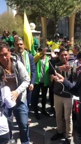 A group of people is gathered outside, with some appearing to be parading or celebrating. In the foreground, a man in a grey Under Armour jacket is seen. Behind him, a man in a suit is waving, adorned with a green and yellow scarf that says "ASFI" on it. There's a building in the background, and several individuals are either carrying flags or wearing green and yellow attire. A drummer is also visible, adding to the festive atmosphere. The weather appears to be sunny and bright.