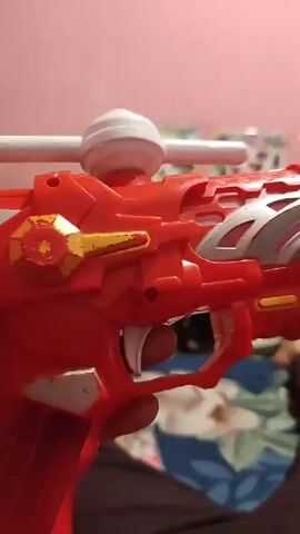 The video is a close-up, shaky, point-of-view shot of a person holding a red and gold toy gun. The gun has a white circular dome on top and a white trigger. The person's thumb is visible pressing down on the trigger. The background is blurry, showing a pink wall and what appears to be a bedspread with floral patterns. The lighting is dim.