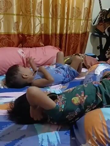 Two children are lying on a bed. The child in the foreground is wearing a green shirt with a character print and is lying on their back with their hands covering their face. The child in the background is wearing a blue shirt and shorts and is lying on their back with their arms and legs extended. A pink bolster pillow is situated between the two children. The bed has a blue and white striped sheet. In the background, there are yellow and brown curtains with a rose pattern. There is also a partially visible black object, possibly a stand or furniture, to the right of the frame. The lighting suggests it is daytime or an indoor setting with artificial light.