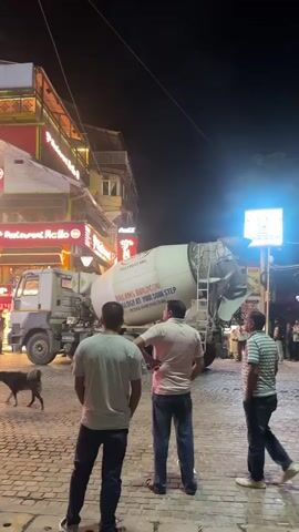 The video shows a nighttime street scene with a cement mixer truck parked on a cobblestone road. Three men are standing in the foreground, facing away from the camera, observing the truck. The truck's drum is tilted, suggesting it is either dispensing or receiving material. Several buildings with illuminated signs, possibly restaurants, are visible in the background. Lights from the buildings and street lamps illuminate the scene. A dog is visible on the left side of the frame. Other people are present in the background, walking and standing. The overall atmosphere appears to be that of an active urban street despite the late hour.