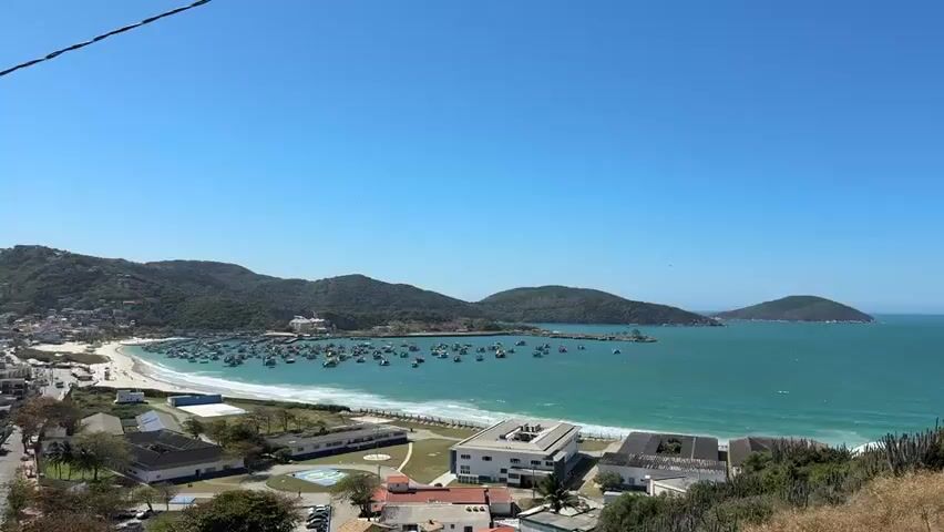 This video shows a panoramic view of a coastal town and bay. In the foreground, there are buildings and vegetation on a hillside. Moving towards the middle ground, there is a sandy beach and a body of water with numerous boats anchored. The boats appear to be various sizes and colors, some with awnings or structures on deck. In the background, there are lush green hills or mountains under a clear blue sky. The water is a vibrant turquoise color. The overall scene depicts a sunny day in a seaside location with a prominent harbor or bay filled with boats.