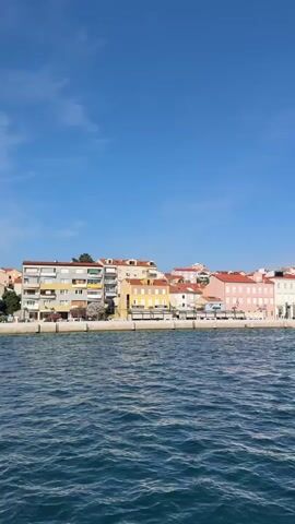 This media file captures a panoramic view of a coastal town under a bright, clear blue sky. The foreground is dominated by the deep blue, choppy water of a bay, with small waves rolling towards the shore.

The midground reveals a picturesque waterfront lined with a vibrant array of buildings. These structures are characterized by their colorful facades, featuring shades of yellow, pink, white, and cream, with terracotta-tiled roofs. The architecture suggests a Mediterranean style, with multiple stories and windows. Palm trees are interspersed along the waterfront, adding to the tropical feel. Several boats and yachts are docked along the harbor, indicating this is a working port or marina.

In the background, the town extends up a gentle slope, with more buildings and a church steeple visible on the higher ground. The overall scene depicts a lively coastal community on a sunny day. The clear blue sky and the bright colors of the buildings suggest it is daytime, likely late morning or early afternoon. The overall mood is serene and inviting, evoking a sense of holiday and relaxation. There is no visible text in the media file. The location is identified as Mali Lošinj, Croatia.