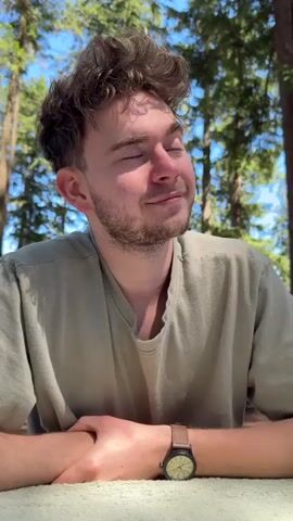 A young man with curly brown hair and stubble sits outdoors, looking at the camera with a slight smile. He is wearing a light olive-colored t-shirt and a watch with a brown band on his left wrist. His hands are clasped in front of him on a textured surface, possibly a blanket or tablecloth. In the background, tall trees with green foliage and a blue sky are visible. The man turns his head to his left, looking down and to the side, before returning his gaze to the camera and smiling again. The scene appears to be set in a park or forest area on a sunny day.