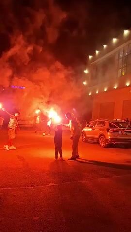 The video shows a nighttime scene with several people holding up red flares, illuminating the area with a strong orange glow. In the foreground, a young child is standing between two adults, one adult is holding a flare up and the other adult appears to be interacting with the child. In the background, there are several other people, some of whom are also holding flares. A building with illuminated windows is visible on the right side of the frame, and a car is parked nearby. The sky above is dark with occasional small fireworks or sparks visible. The ground is paved, and the overall atmosphere appears festive or celebratory.