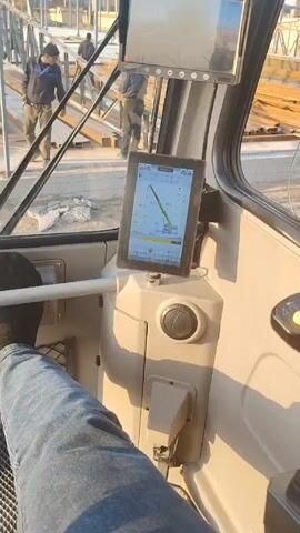 The video shows the perspective of a crane operator inside the cab of a crane. The operator has their leg up on a support, and a tablet is attached to the dashboard with crane operation details. Through the front window, workers on the ground are visible moving metal beams and trusses in a construction area. The crane hook and cable are visible overhead, presumably controlled by the operator. There are steel posts standing and the beginning structure of a building going up.