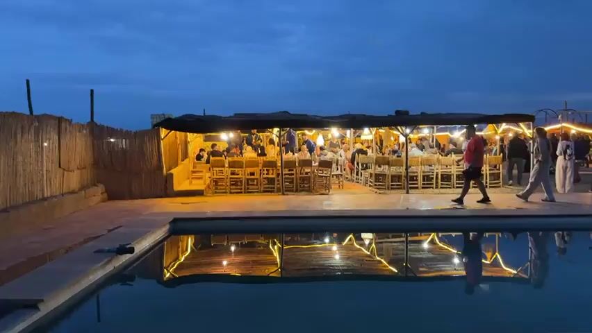 The image depicts an evening scene at dusk or early night in Sidi Badhaj, Morocco. In the foreground, a swimming pool with dark, calm water reflects the warm, golden light emanating from an adjacent open-sided structure.

This structure, resembling a large, elongated tent or shelter with a dark roof, is brightly illuminated by multiple bare light bulbs strung overhead. Inside, a group of approximately 20-30 people, primarily adults, are seated at long, rustic wooden tables on high wooden chairs, engaged in a social gathering or dinner. The atmosphere appears lively and celebratory.

At the center of the structure, entertainment is being provided. One man in a blue traditional robe stands, holding what appears to be a small, decorated vessel, while another man to his left plays a large drum. Several seated guests are turned towards them, some clapping, indicating an engaging live performance.

Outside the structure, a man in a red shirt walks along the paved area between the structure and the pool, and another person in light-colored clothing walks further to the right. To the left of the structure, a rustic fence made of reeds or bamboo is visible. The sky above is a deep blue, indicative of twilight. No visible text is present. The overall scene suggests a cultural dining and entertainment experience.